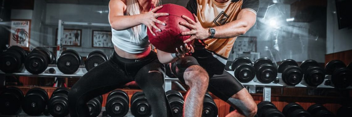 A man and woman perform a tandem workout with a medicine ball in a gym setting.